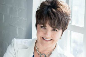 Professional headshot of Deborah Bass, smiling, wearing a white blazer and necklace, seated by a window with natural light. 