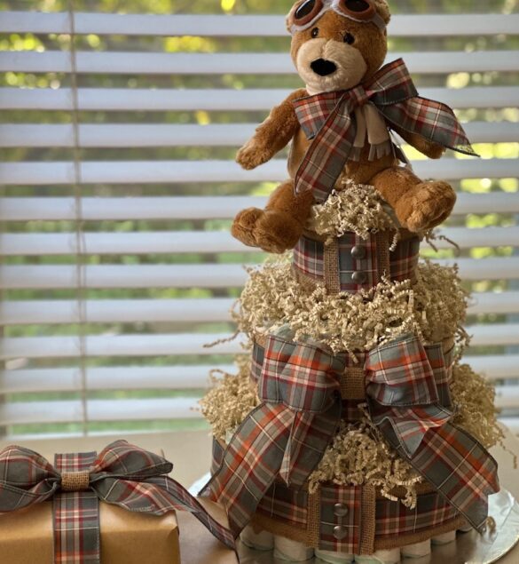 three-tiered diaper cake topped with a plush teddy bear wearing aviator goggles, wrapped in brown and cream ribbons, displayed on a table with window blinds in the background