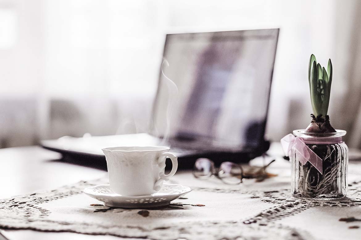 A steaming hot cup of tea on a desk with an open laptop and pink flowers