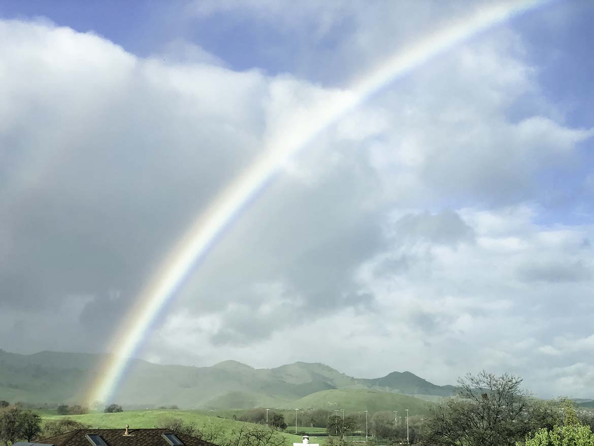 A stunning rainbow shoots from the green hills amid cloudy skies—a view from my bedroom window