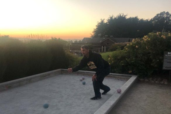 A man is in mid-throw playing bocce ball with a glorious sunset behind him