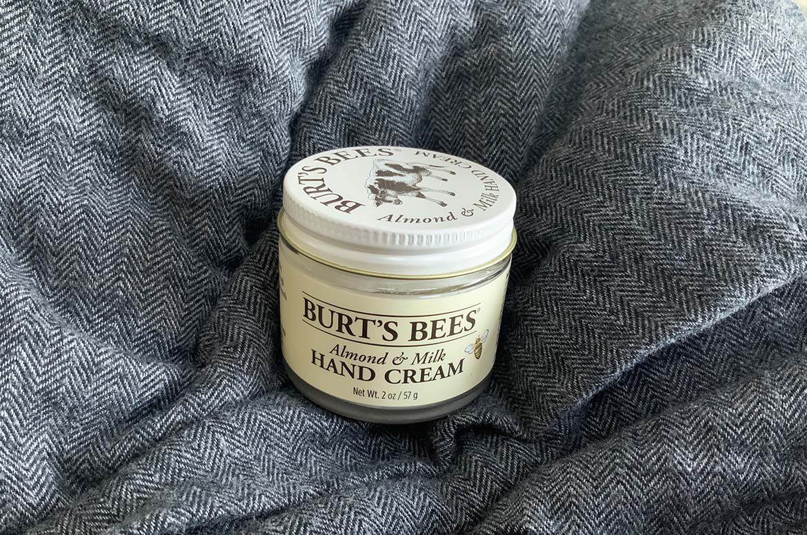 A jar of Burt's Bees Almond & Milk Hand Cream rests on a fluffy gray blanket