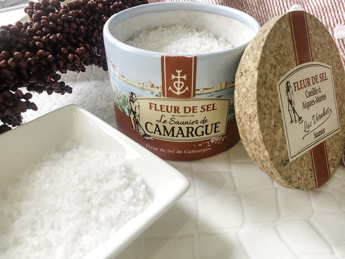 A box of Fleur de Sel (salt) sits open with its cork lid leaning against it. Next to is a white bowl filled with sparkly Fleur de sel.
