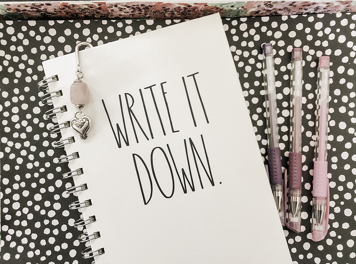 A spiral bound notebook titled Write It Down lays on a black and white polka dot paper with a jeweled and silver bookmark in it. Colorful gel pens lay at its side.