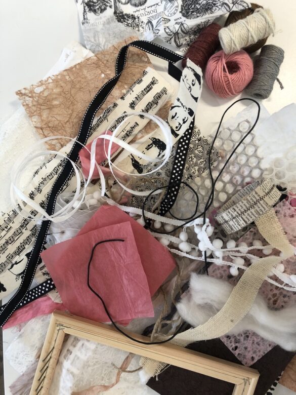 Various colorful ribbons and papers in white, creams, pinks, and black lay jumbled on a table next to a handmade loom.