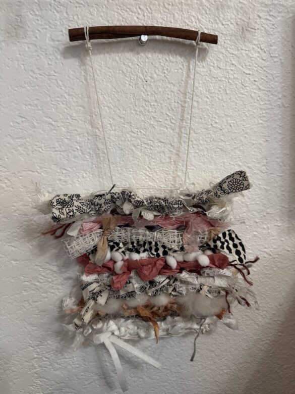 A woven piece of art made from ribbons and papers hangs on a wall.