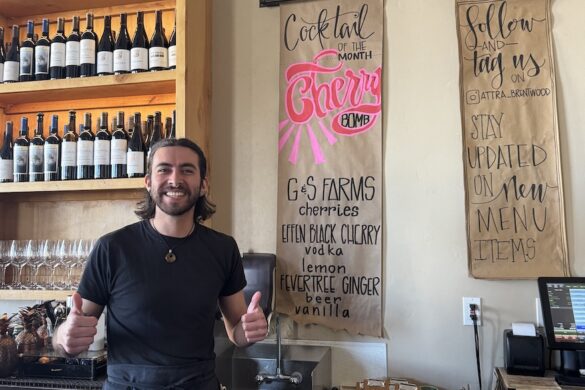 A server in a restaurant holds two thumbs up and smiles big next to the cocktail of the month sign: Cherry Bomb at the Attraversiamo Restaurant in Brentwood, California
