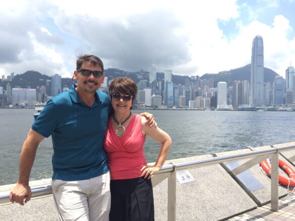 A happy couple, the man with his arm around his wife, on a boat with the Hong Kong skyline in the background