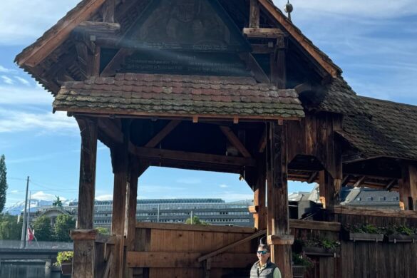 Man standing at the wooden entrance of Lucerne’s Chapel Bridge, with timber beams, a tiled roof, and painted triangular panels above