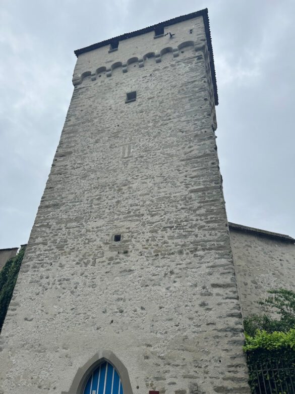 View looking up at Schirmer Tower along Lucerne’s Musegg Wall, with a tall stone façade, arrow slits, and a blue-barred arched doorway