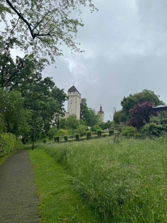 Pathway leading through a lush green park toward two medieval towers of Lucerne’s Musegg Wall on a rainy day