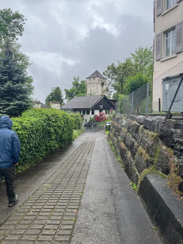 Rainy path leading through a quiet Lucerne neighborhood toward a stone Musegg Wall tower, with a person walking ahead in a blue jacket