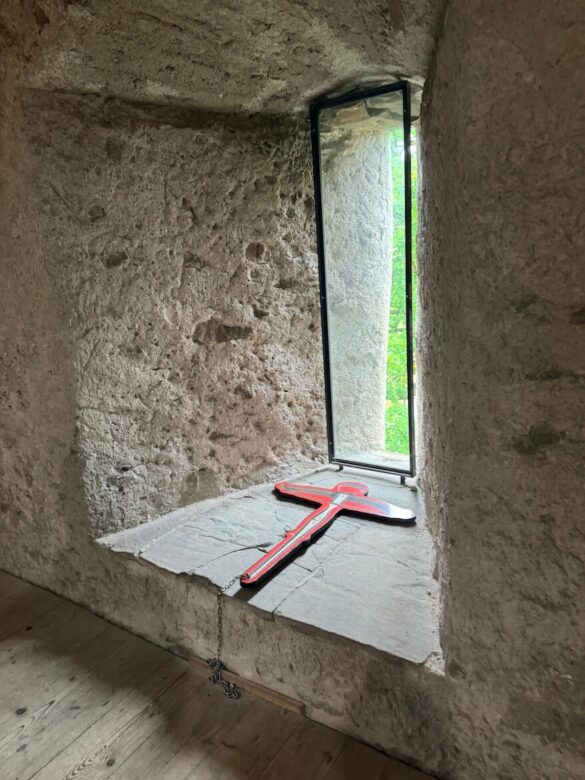 Narrow medieval stone window with an interpretive red bow-shaped sign, showing how archers used the opening to defend Lucerne’s Musegg Wall