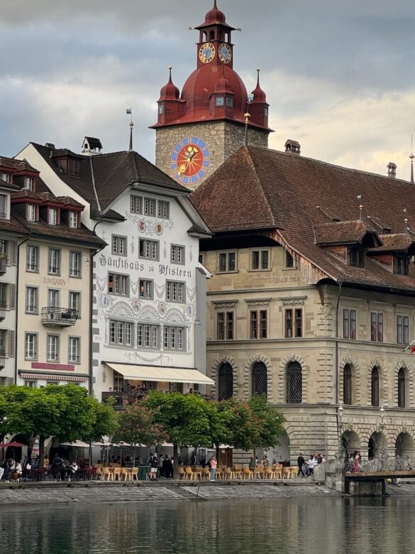 Historic buildings along the Reuss River in Lucerne, including a red-roofed clocktower with an astronomical dial, viewed under cloud cover