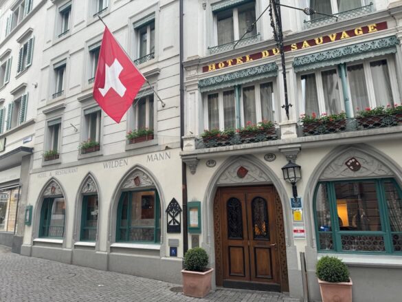 Exterior of Hotel Wilden Mann in Lucerne with Gothic-style arches, Swiss flag, and green window shutters and flower boxes