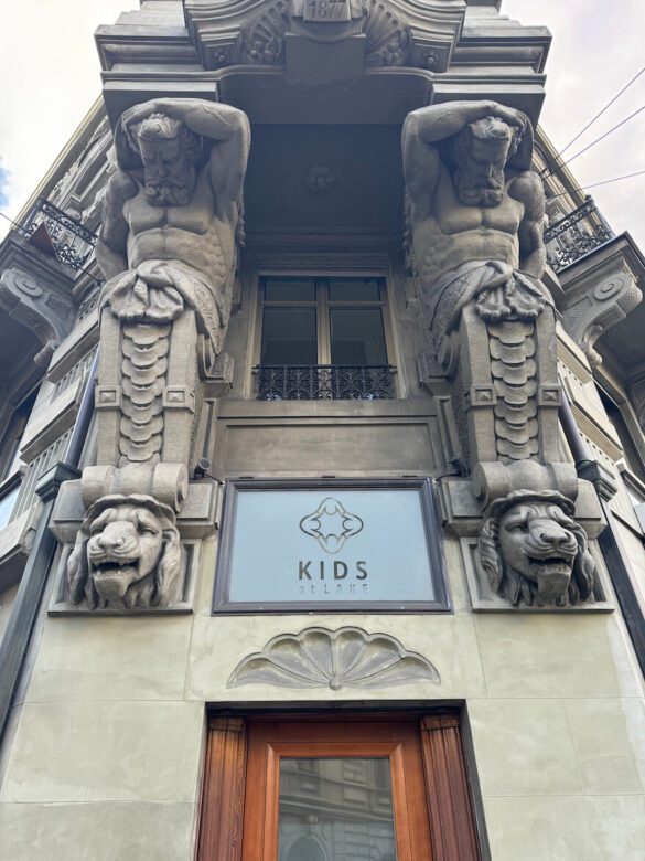 Stone building in Lucerne with carved male atlantes and lion heads above a modern “Kids at Lake” sign