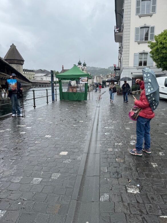 Rainy scene along the Reuss River in Lucerne with Chapel Bridge on the left, a market tent in the center, and pedestrians walking along wet cobblestones