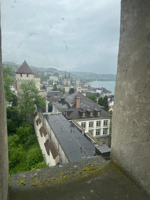 View of Lucerne on a rainy day seen through a window slit in the Musegg Wall, with stone towers, rooftops, and twin cathedral spires visible in the mist beyond.