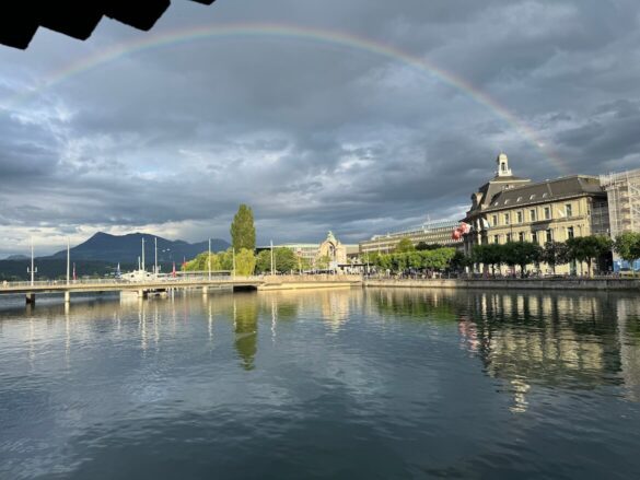Rainbow arcs over Lucerne’s riverfront with reflections in the water, historic buildings, and Mount Pilatus in the background.