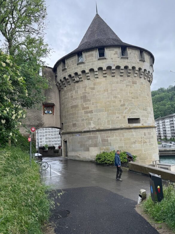Stone round tower with battlements in Lucerne on a rainy day, viewed from street level.