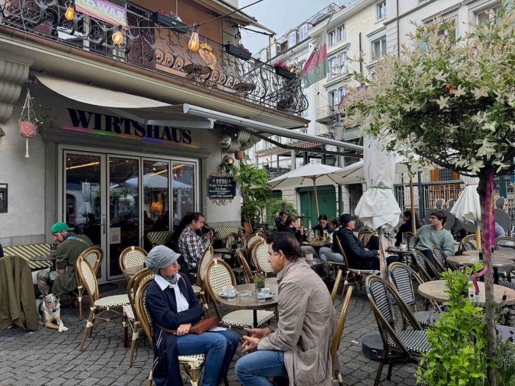 Outdoor dining area of Wirtshaus Krienbrüggli restaurant in Lucerne, with people seated at tables under umbrellas, wicker chairs, flower pots, and a lively atmosphere on a cloudy day.