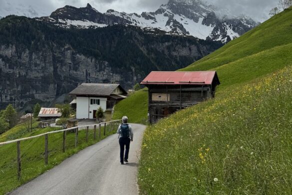 Woman with backpack walking up a paved path toward wooden barns, snowcapped mountains and dark clouds ahead