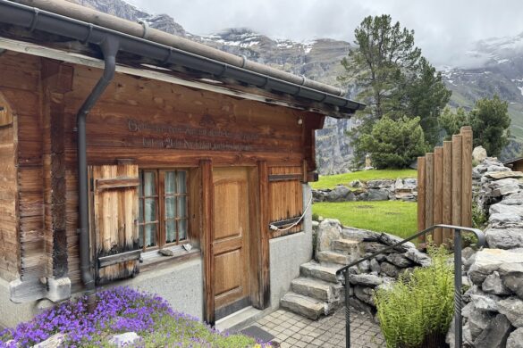 Wooden chalet with flower beds and faint engraving above the doorway, framed by alpine stonework and purple blossoms