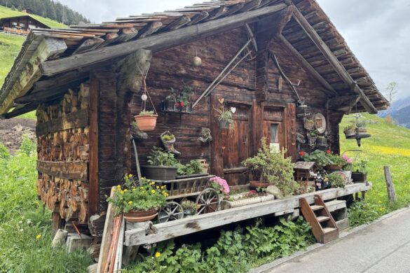 Weathered wooden chalet front with log pile walls, flowerpots, and wagon cart filled with blooms