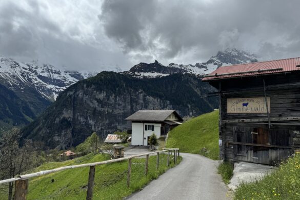 Small wooden barn with sign reading “Willkommen in Gimmelwald,” houses and snowcapped mountains behind