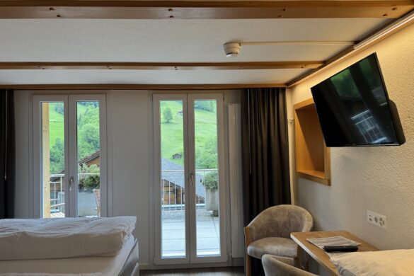 Hotel room interior with twin beds, chairs, desk, and wall-mounted TV, with glass doors opening to a balcony and hillside view