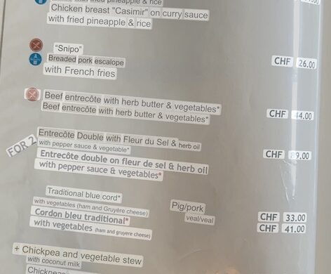 Smartphone screen showing a restaurant menu translated from German to English with Google Lens; items include spaghetti Bolognese, chicken curry, beef entrecôte, cordon bleu, and chickpea vegetable stew.