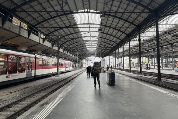 Ron walking with a large backpack through the wide platform at Lucerne train station, with a red and white train on the left and arched roof overhead