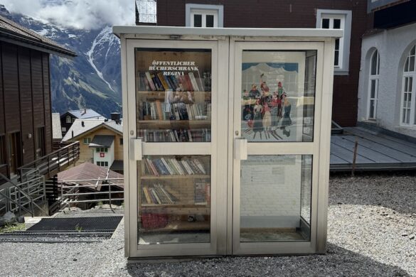Outdoor book-exchange cabinet in Mürren with shelves of books inside glass doors, Hotel Regina and mountain backdrop behind
