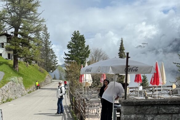 Woman in a white jacket standing on a paved street in Mürren with a stone wall café terrace, umbrellas, and misty clouds in the background