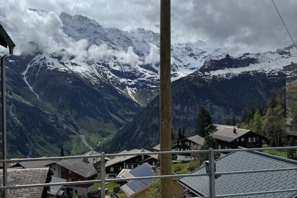 View over Mürren rooftops with solar panels toward a deep valley and snow-covered peaks under dramatic clouds