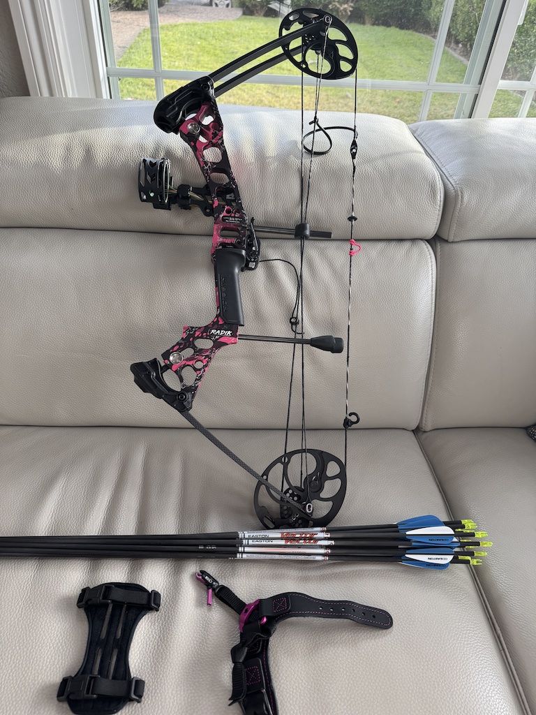 Black and red compound bow resting on a beige couch with a quiver of arrows beside it