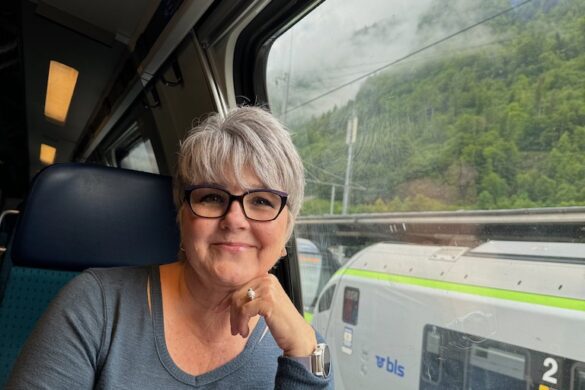 Deborah with wet hair sitting by a train window en route from Lauterbrunnen to Basel, Switzerland, with green hills and reflections visible outside