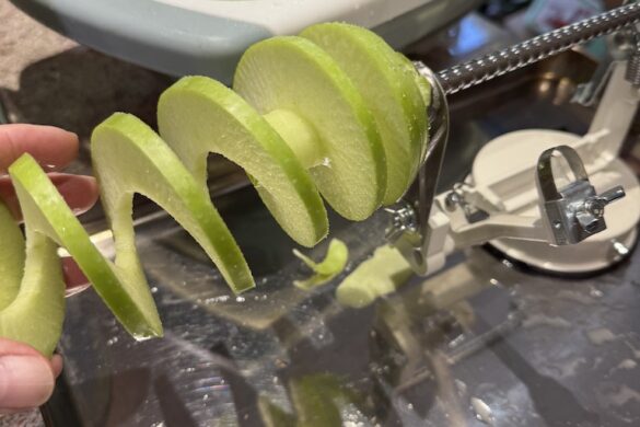 Spiral-sliced green apple on a hand-crank peeler beside a bowl of whole apples