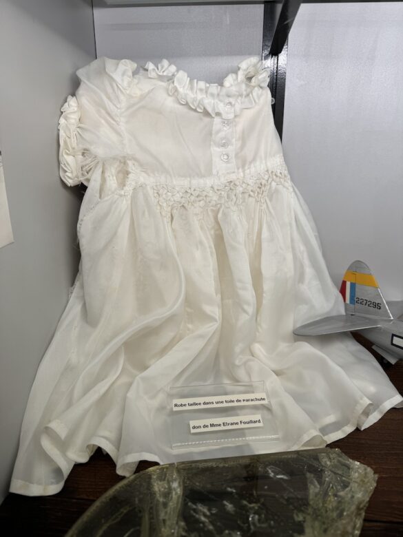 Small white baby dress displayed in a museum case, made from WWII parachute silk.
