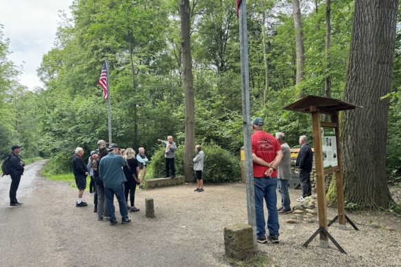 Tour guide standing before visitors near flagpoles at the Audie Murphy Memorial in France, demonstrating the direction of air support.