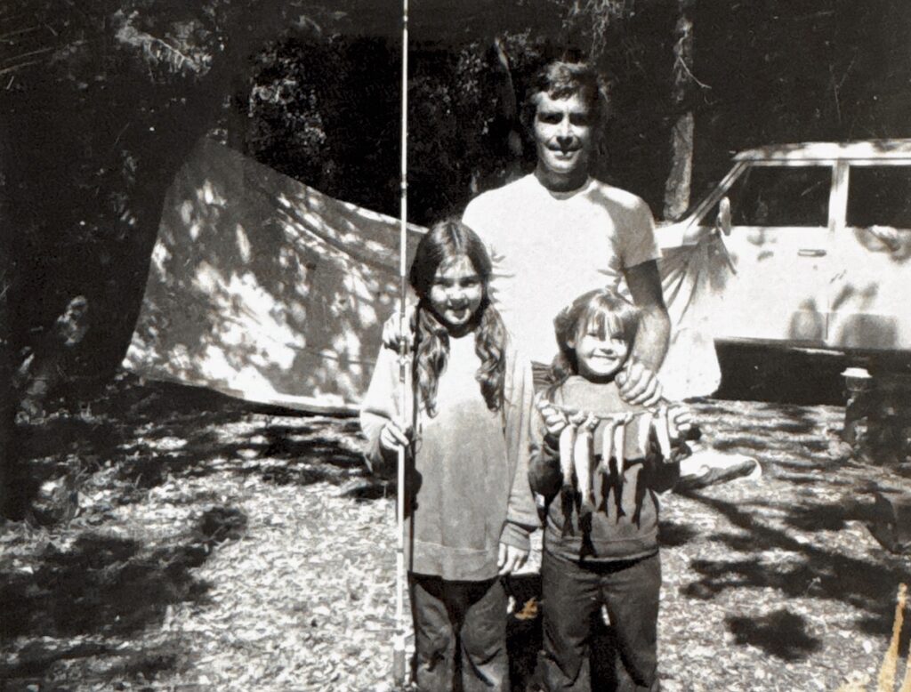 black and white photo of young Deborah and her sister holding fish beside their father near a stream