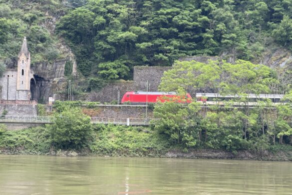 Train moving along hillside beside the Rhine River near small castle-style façade by a railway tunnel