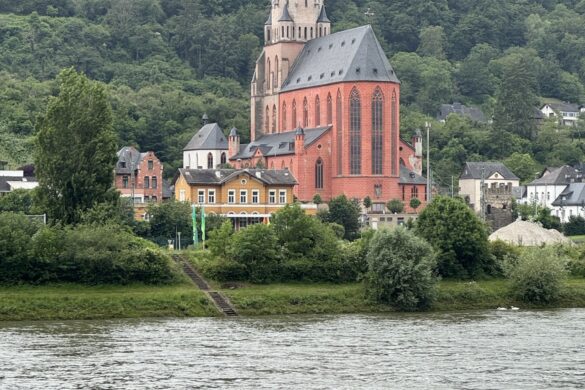 Red brick church with tall spire beside the Rhine River with green hillside behind