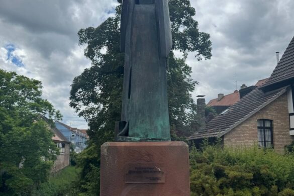 abstract bronze sculpture of a standing figure on a stone bridge pedestal with water and leafy green background under cloudy sky