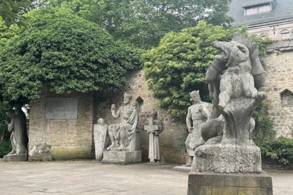 Weathered stone statues of historical figures standing along a brick wall beneath trees near Speyer Cathedral