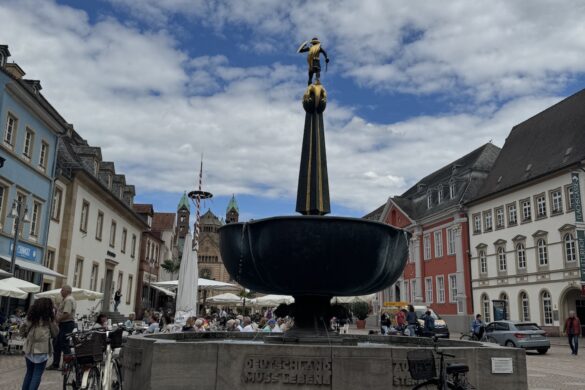 large stone fountain with bronze figure in a busy town square with colorful buildings and cathedral towers behind