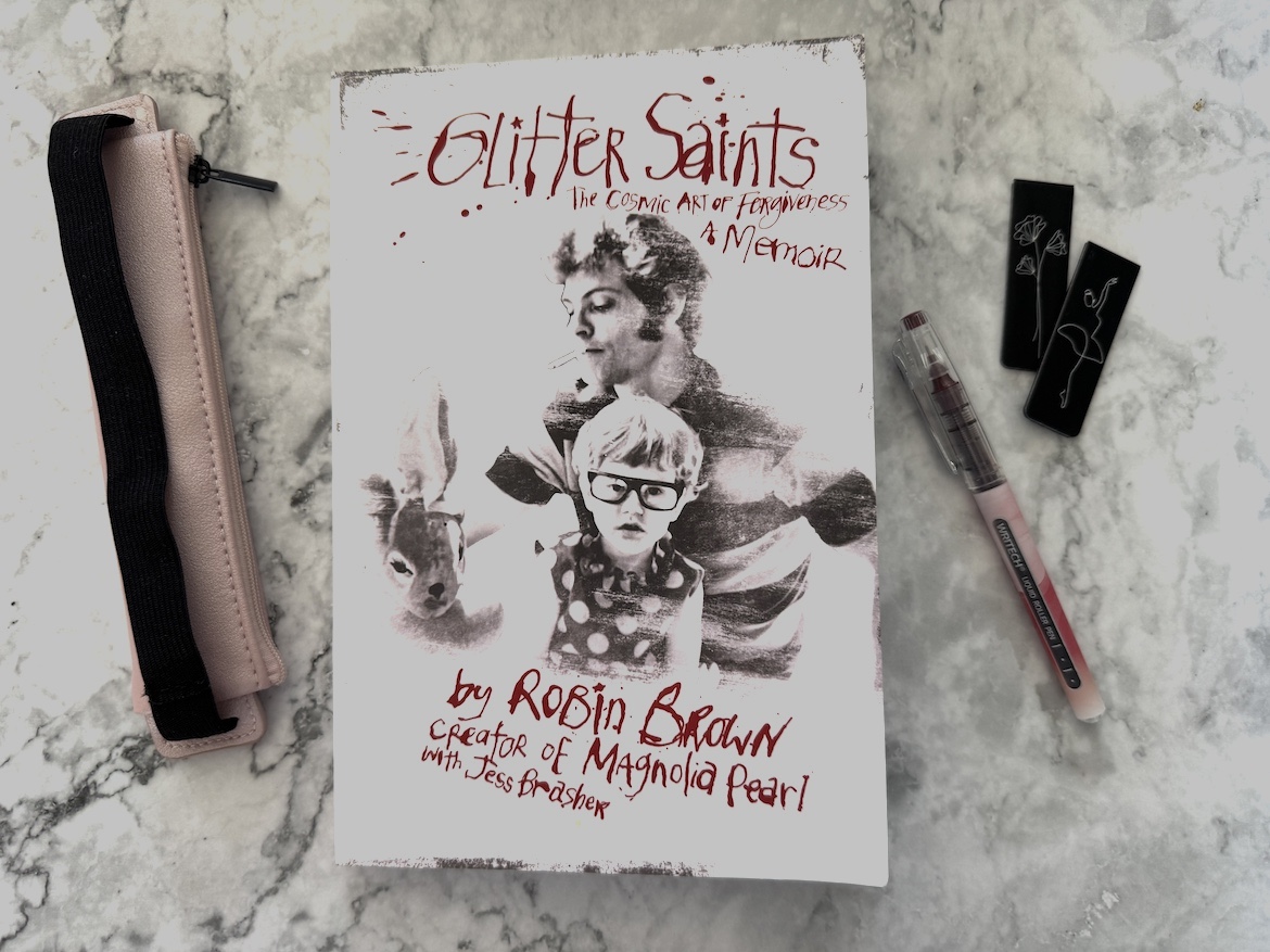 book Glitter Saints The Cosmic Art of Forgiveness by Robin Brown resting on a light surface with bookmark and pen nearby