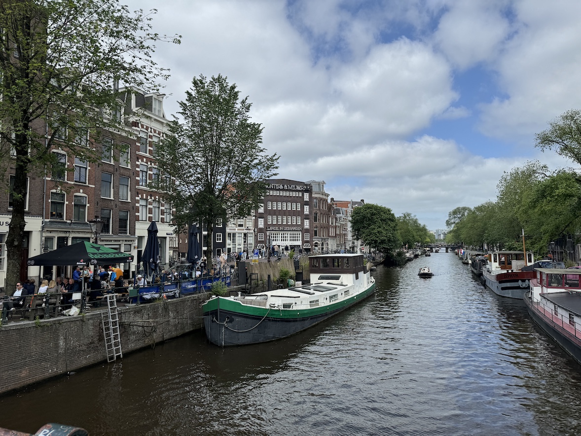 wide amsterdam canal with boats, brick buildings, trees, and a bridge under a partly cloudy sky
