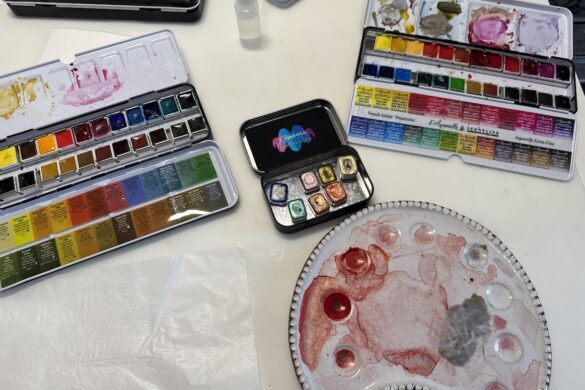art workspace with watercolor palettes and chroma metallic paint tins in the center alongside mixing palette and supplies