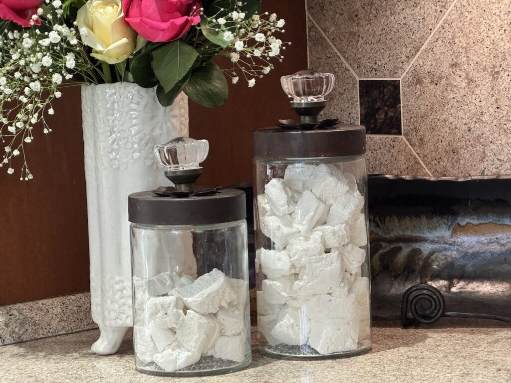 Homemade marshmallows in glass jars on a kitchen counter with flowers in the background